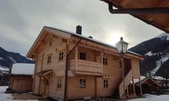 A cozy wooden house in a snowy landscape. Mountains can be seen in the background and the sky is overcast.