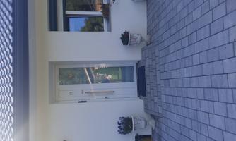 An entrance to a modern house with a white wall.  
In front of the door, there are two pots with plants and the ground is paved with gray stones.