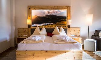 A cozy bedroom with a large bed and wooden furniture. In the background hangs a picture of a landscape with the sun.