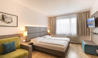 A modern hotel room with a double bed and a cozy seating area. Large windows provide plenty of natural light.