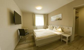 A bright room with a double bed and a television. There is a chair and a small table next to the bed as well as a wardrobe.