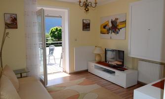 A bright room with a white sofa and a television. The door leads to an outdoor area with chairs and a green view.