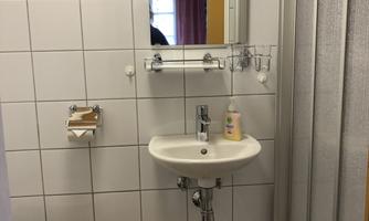 A small bathroom with a sink, a mirror, and a shower. The walls are tiled and there is a toilet as well as a small table.