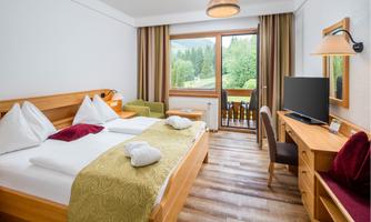 A cozy bedroom with a large bed, wooden furniture, and a desk. The window offers a view of the green landscape outside.