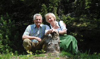 An older couple is smiling while sitting in the forest next to a tree stump. In the background, green trees and nature are visible.