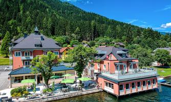 A picturesque hotel by the shore of a lake, surrounded by green mountains. The terrace is equipped with tables and umbrellas, perfect for relaxing.