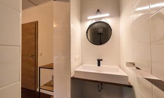 A modern bathroom with a white sink and a round mirror. The walls are light-tiled, and there is a wooden cut floor.