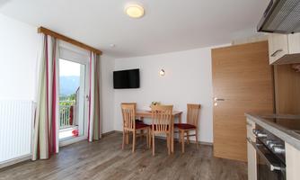 A modern living kitchen with a dining table and chairs. A window with a view of the mountains and a well-equipped kitchenette are also visible.