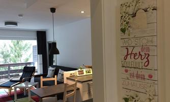 A cozy living area with a modern sofa and a dining table. On the wall hangs a decorative sign that reads "Where your heart is, feels like home."