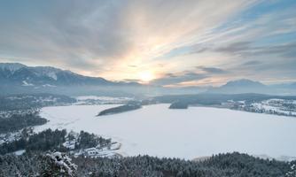 A peaceful lake in winter with a snow-covered landscape. The sky is cloudy, but the sun breaks through the clouds.