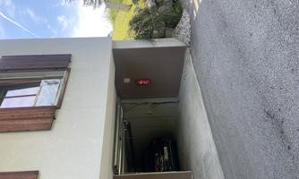 A garage under a residential building with a car inside. In the background, there are trees and a blue sky visible.