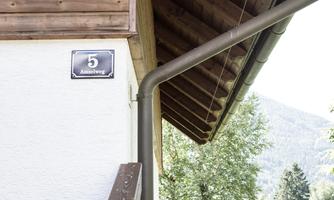 A part of a house with wooden siding and a house number 5. In the background, trees and a mountain landscape can be seen.