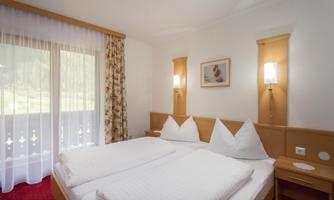 A cozy room with a double bed and white bed covers. Large windows with curtains provide a view outside.