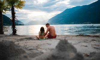 A man and a girl are sitting on the beach, looking at the lake. In the background, mountains and a cloudy sky are visible.