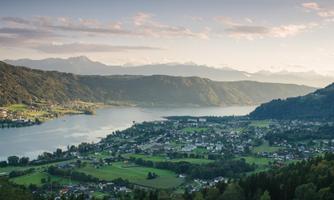 A picturesque landscape with a calm lake and gentle hills. Small houses are scattered, and the mountains frame the scene.