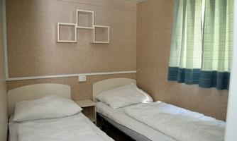 A simple bedroom with two single beds and neutral walls. A window with green curtains brings daylight into the room.