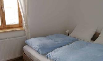 A bright bedroom with a large bed and blue blankets. A window with curtains and a simple lampshade complement the room.