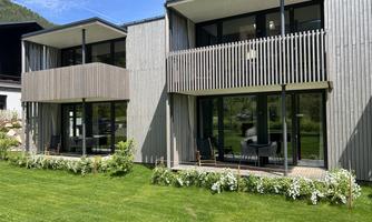 Modern wooden buildings with large windows stand in a green garden. The surroundings are sunny and peaceful.
