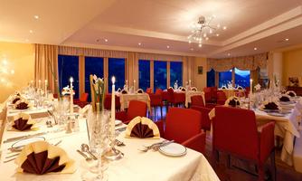 An elegant restaurant with festively set tables and red chairs. Large windows offer a view of the evening atmosphere.