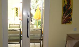 A bright interior with a table and a door leading to a garden. In the background, chairs and a sun umbrella are visible.