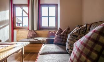 A cozy living space with a wooden sofa and decorative pillows. The windows let in plenty of daylight and offer a view outside.
