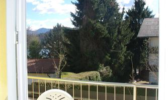 A balcony with a round table and two chairs. In the background, green trees and a clear sky are visible.