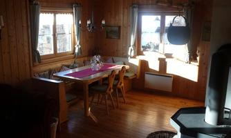 A cozy dining area in a wooden house with a large table and chairs. Sunlight is streaming through the windows and there is an inviting atmosphere.