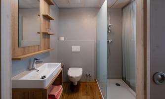 A modern bathroom with wooden shelves, a sink, and a shower. The walls are bright, and the wooden floor provides a warm atmosphere.