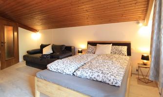 A cozy bedroom with a double bed and a seating area. The wooden ceiling and soft lighting create an inviting atmosphere.