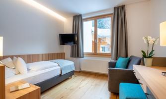 A modern hotel room with a single bed, a sofa, and a desk. Large windows provide plenty of daylight.