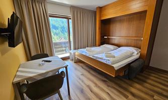 A cozy hotel room with two beds, a table, and a chair. Large windows offer a view of the mountains.