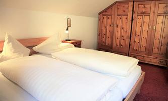 A cozy bedroom with two single beds and white bedspreads. In the background, there is a wood-colored wardrobe, and a bedside lamp provides soft light.