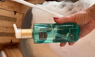 A hand is holding a bottle of shower gel in an elegant design. In the background, a bathtub with foam is visible.