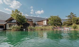 A tranquil lake with clear, greenish water and surrounding trees. In the background, several buildings create a relaxed atmosphere.