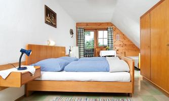 A cozy bedroom with a double bed and blue bedspreads. There is a desk with a lamp and a view of the outside area through a window.