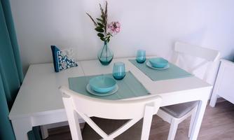 A cozy dining area with a white table and two chairs. On the table are blue glasses and dishes, as well as a vase with a flower.