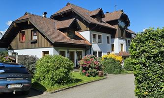 A large, charming house surrounded by colorful flowers and green shrubs. A car is parked in the driveway, and the sky is clear and blue.