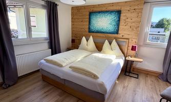 A cozy bedroom with a large bed and wooden paneling on the wall. It has large windows that let in plenty of light.