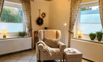 A cozy living space with a comfortable armchair and a small table. The windows let in plenty of daylight, and the decor creates an inviting atmosphere.