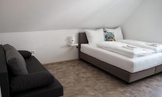 A simple and modern bedroom with a double bed and a gray sofa. The room has bright walls and a warm wooden floor.