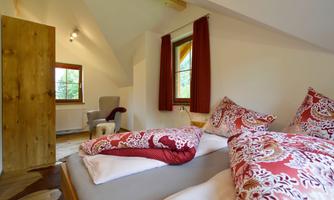 A cozy bedroom with a large bed and decorative pillows. The room has a window with red curtains and a bright wooden ceiling.