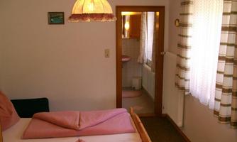 A cozy bedroom with a double bed and pink bedding. The room has a window with curtains and leads to a bathroom.