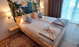 A cozy bedroom with a double bed and decorative pillows. The wall is adorned with a floral pattern, and large windows provide plenty of daylight.