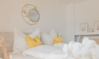A cozy bedroom with a bed decorated with gray and yellow pillows. A round mirror hangs on the wall and there are some framed pictures.