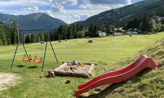 A playground in an idyllic landscape with swings and a slide. In the background, green hills and some houses can be seen.