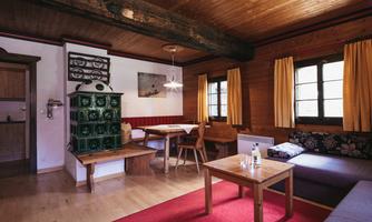 A cozy living room with wooden ceilings and large windows. There is a table with chairs, a couch, and a traditional fireplace.