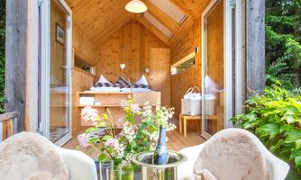 A cozy wooden house with large windows and a view of nature. On the table is a bottle of champagne and fresh flowers.