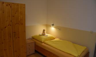 A simple bedroom with a single bed and yellow bedding. Next to the bed is a nightstand and a wooden cupboard against the wall.