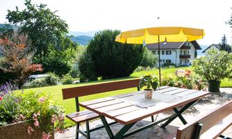 A sunny outdoor area with a wooden table, benches, and a yellow parasol. Surrounded by colorful flowers and green vegetation.