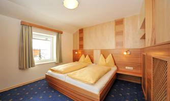 A cozy bedroom with a double bed and two large pillows. The walls are designed in warm wood colors, and the room has a window view outside.